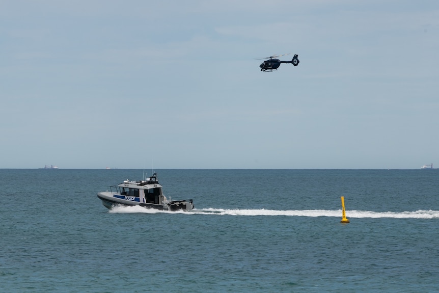 A water police speedboat travels in open ocean near the shore, while a helicopter flies above.