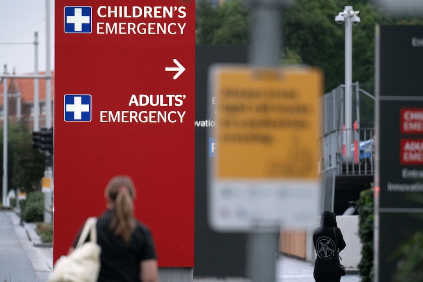 A large red sign on a footpath that says 'Adults' Emergency' with an arrow. Two people are walking past it.