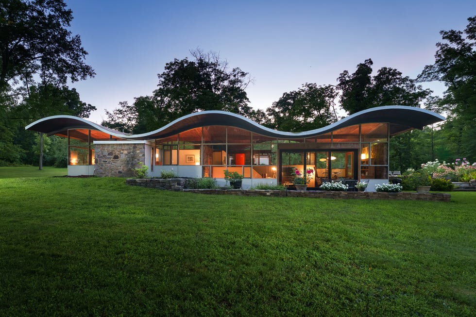 Modern house with curved roof, large windows, and surrounding greenery at dusk.