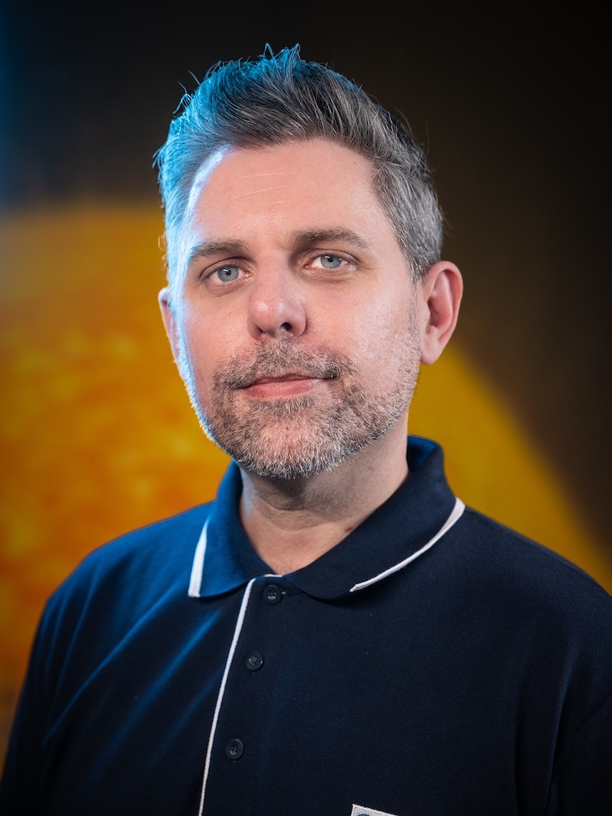 A man in a blue collared shirt standing in front of a yellow and black background.