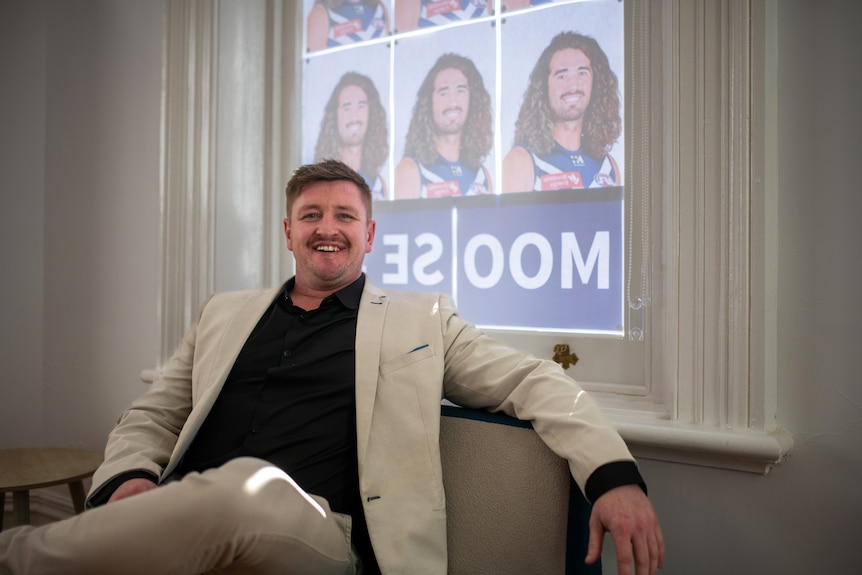 A man smiles in a white suit next to a window covered with posters featuring a football player.