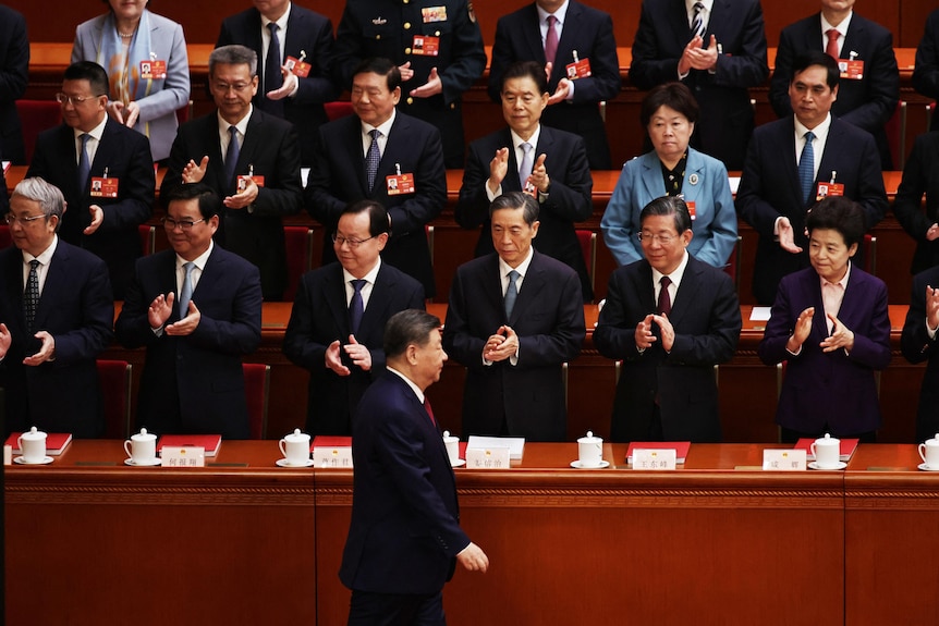 A man in a dark suit and red tie walks past rows of clapping officials.