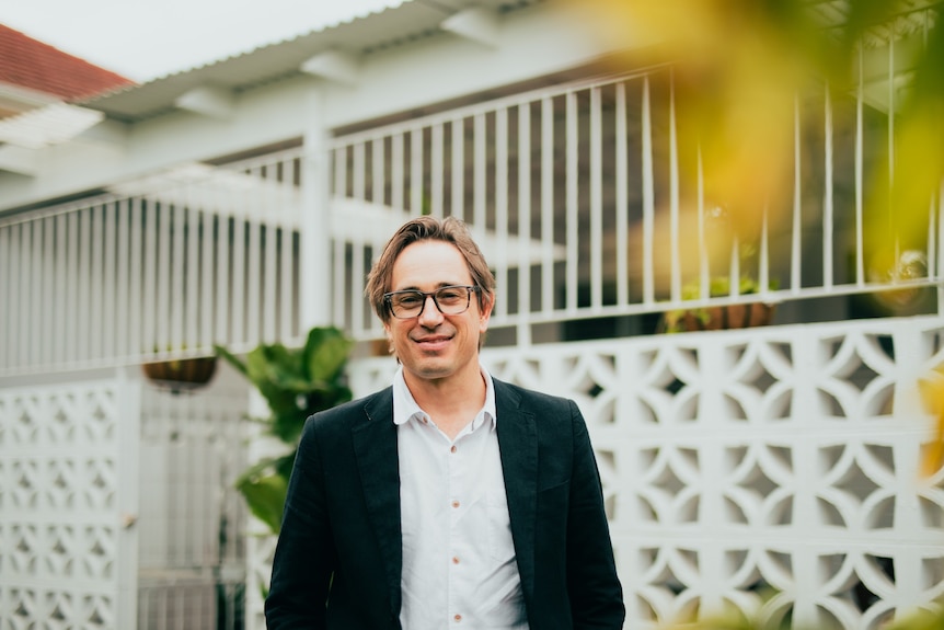 Trent Dalton, a man in his mid-40s, in glasses and blazer, stands smiling outside a suburban home.
