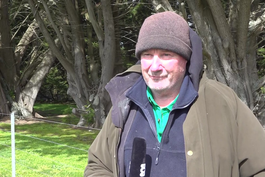 Farmer in beanie and grey jacket standing in front of grass, trees and fencing at his regional property.