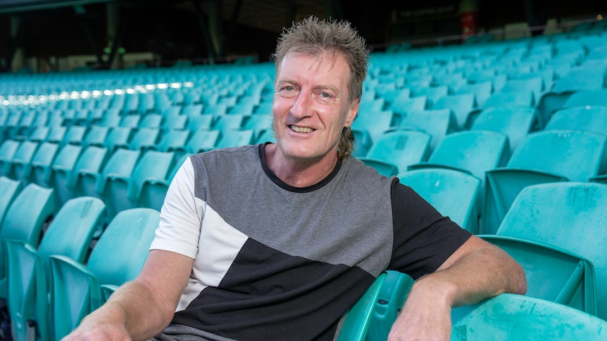 A man sits in a grandstand at the SCG smiling at the camera.