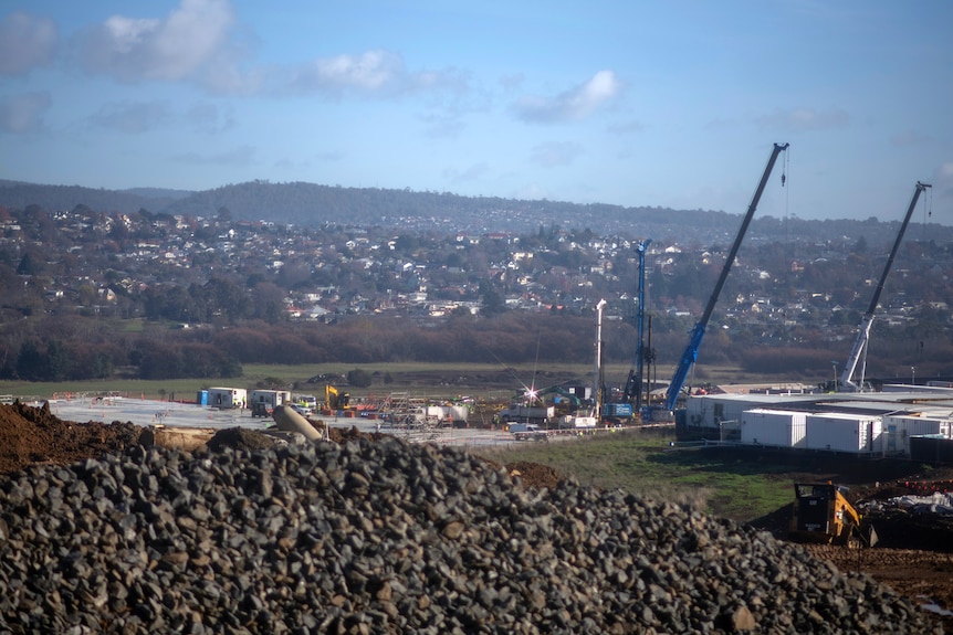Blue cranes at a distant construction site in a valley dotted with homes under a blue hazy sky.