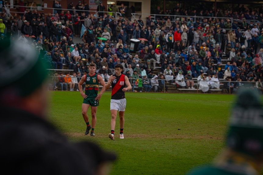 Two players on the field as crowd watches on. One is in green uniform; the other red, black and white.