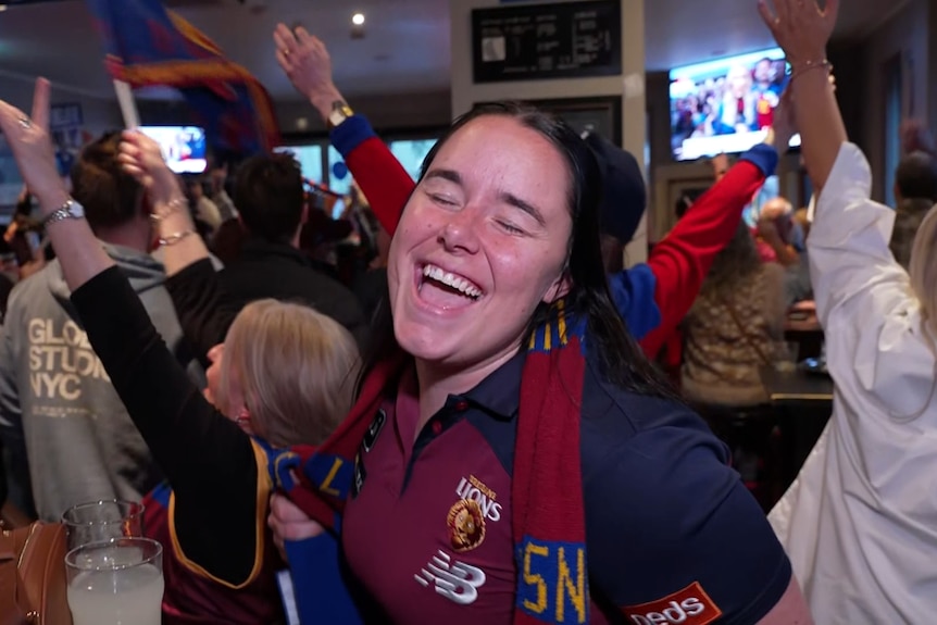 A woman with a Brisbane Lions scarf closes her eyes, singing, as people behind her hold their arms in the air.