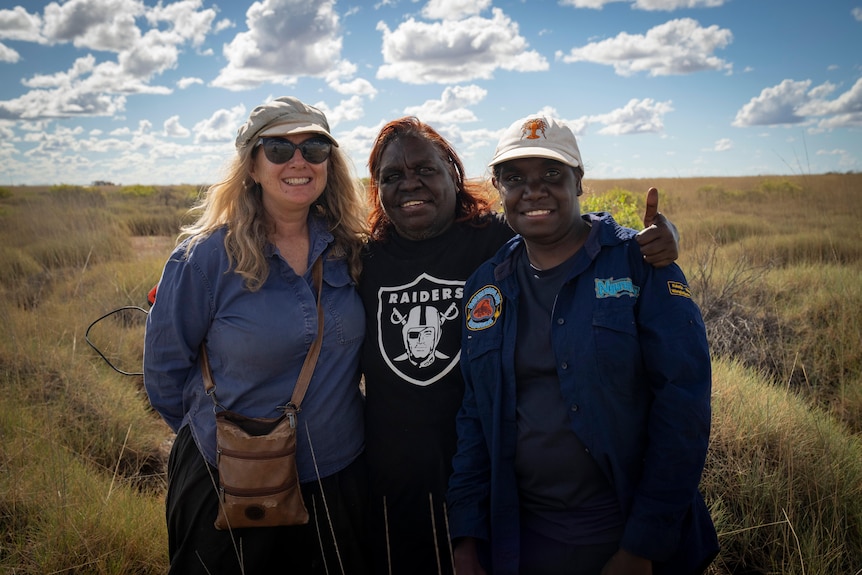 A white woman with sunglasses, a middle-aged Aboriginal woman and a younger Aboriginal woman stand together by spinifex.