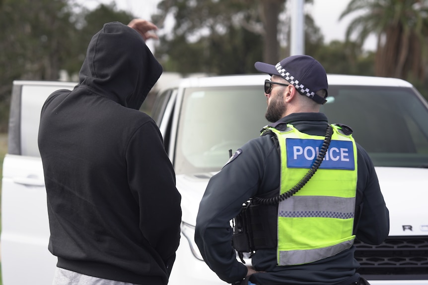 A photo taken from behind a person in a black hoodie talking to a police officer near a white car.