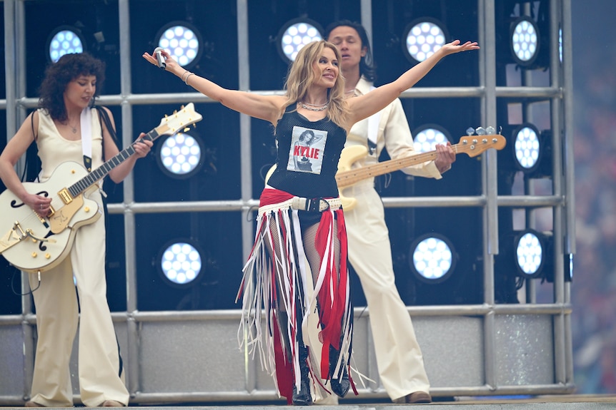 Kylie Minogue wearing a black tank top with her face on it, and a white belt with red, white and black strips