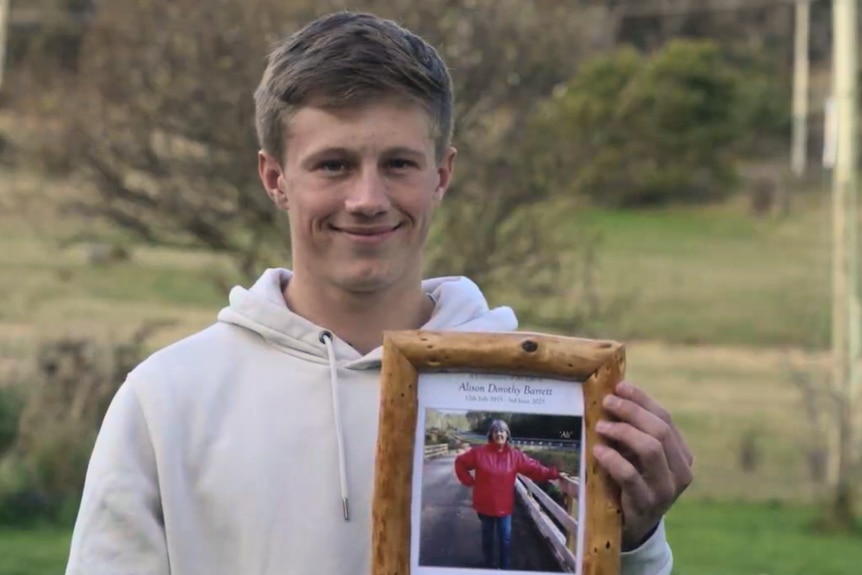 A young man in a white hoodie holding a photo frame of an older woman, smiling at the camera.