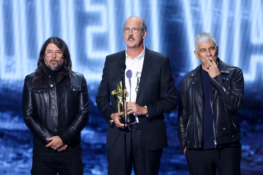 Pat Smear smoking a cigarette in a leather jacket with Dave Grohl also in leather on stage Krist Novoselic holding award