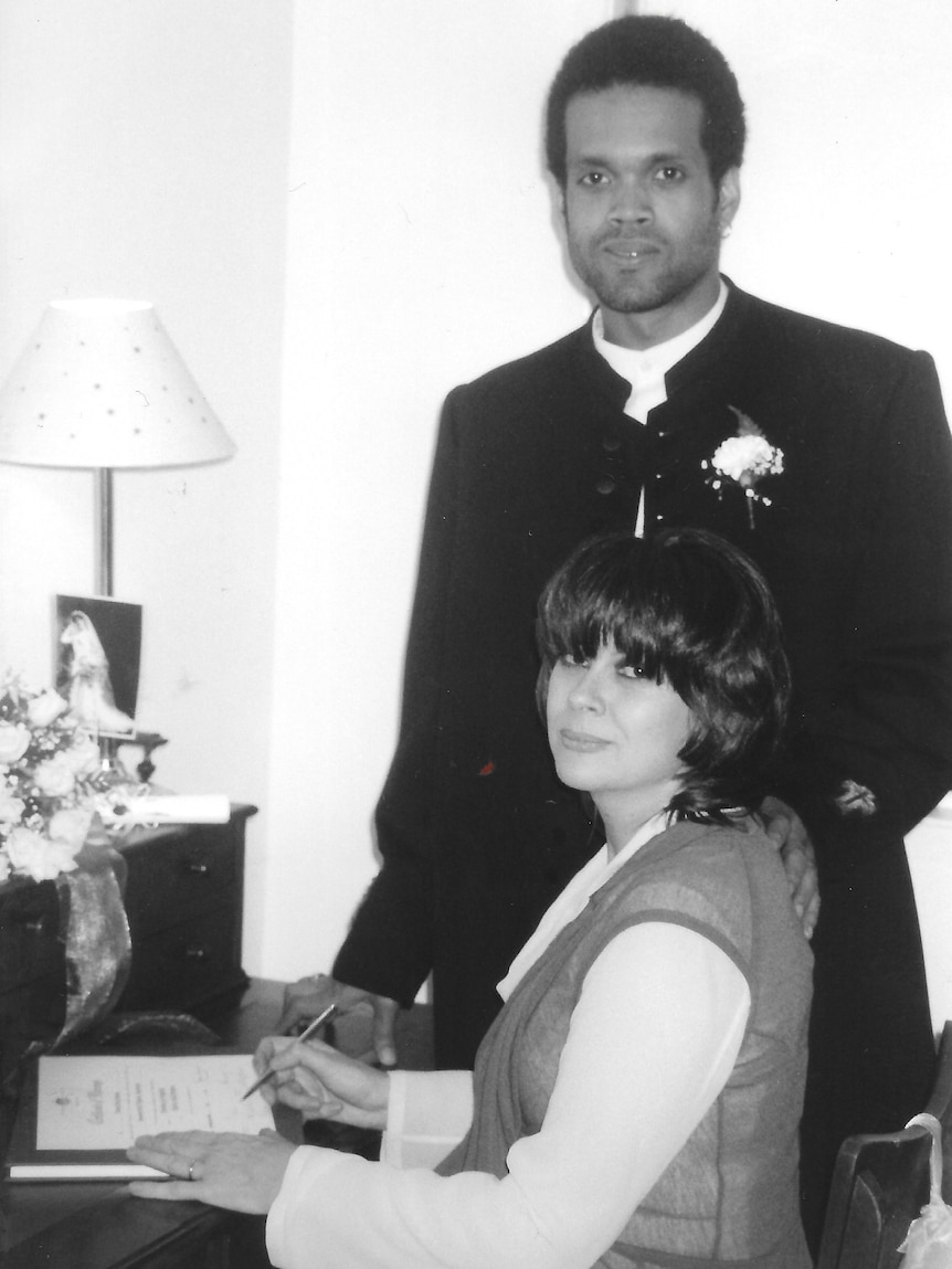 Chrissy sits signing her wedding certificate with Charley behind her in a suit with his hand on her shoulder.