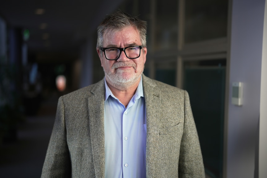 Man with dark rimmed glasses, a blue collared shirt and tweed jacket