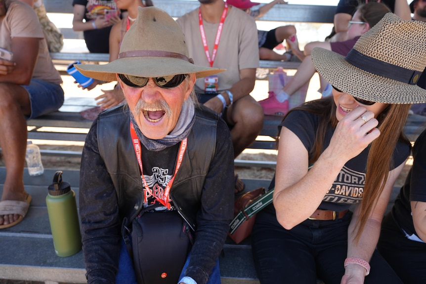 Man sitting in stand wearing Akubra hat looks ahead with sunglasses on and opens mouth