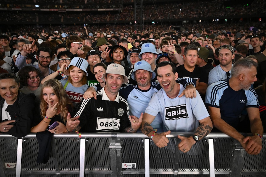 Fans wait at the barricade in front of a stage, cheering in Oasis merchandise.