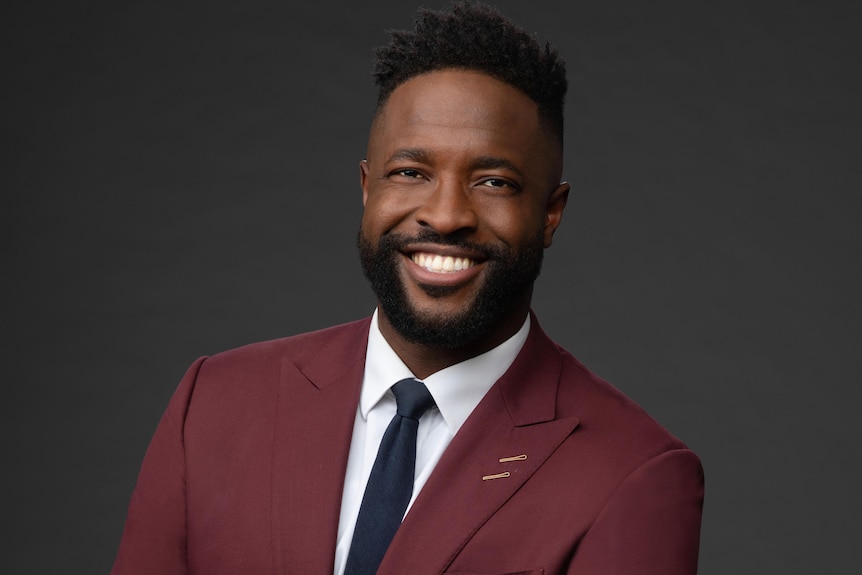 A Black man with a full head of hair, clipped on sides, beard and moustache, maroon suit, navy tie, white shirt, smiling