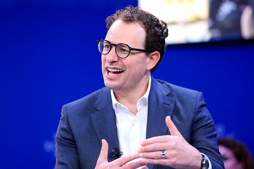 A man in a suit and shirt, with short dark hair and glasses, sits talking in front of a blue background and TV screen.