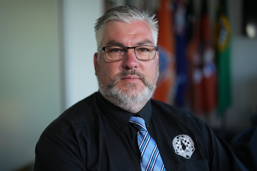 A man in a black collared shirt and blue tie, sitting inside a room and looking serious.