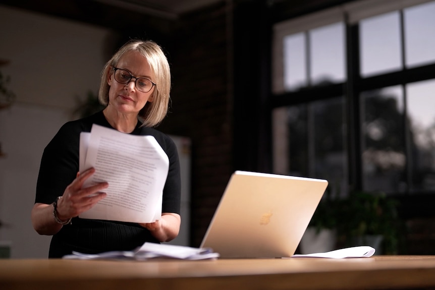 A woman sits at a desk indoors, looking down at pages in her hand as a laptop sits open.