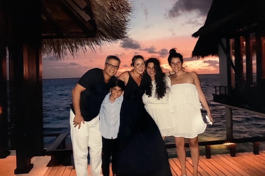 Two adults and three children smile at the camera in what is clearly a tropical, beach-side location.