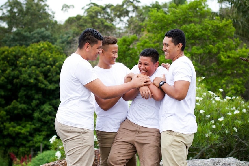 Four young men in white t-shirts with their arms around each other.