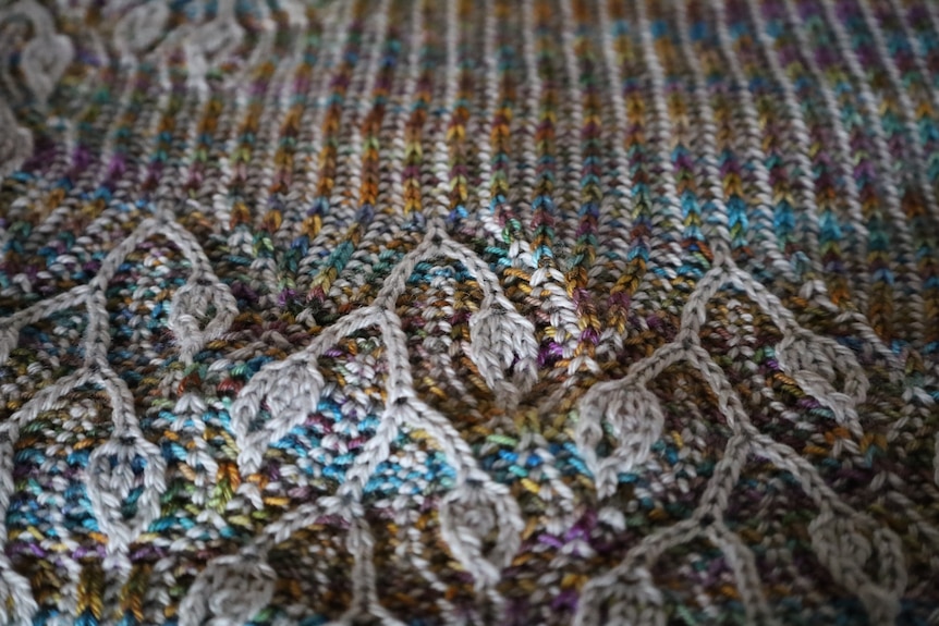 A close up of a handknitted shawl of green, blue, purple and yellow with raised stitches making leaf patterns in grey.