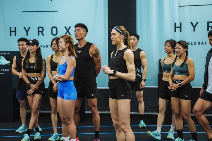 A woman in workout clothes stands listening amid a group of others.