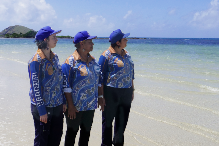 Three women wearing blue collared shirts and caps stand by the sea on a sunny day, looking out at clear waters.