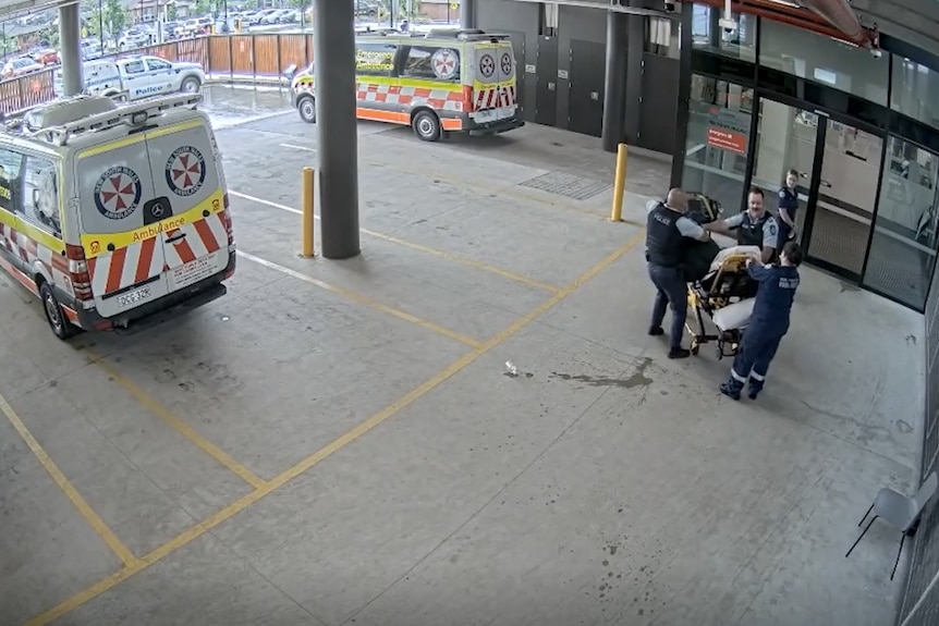 A cctv picture of two ambos to the left, and then two paramedics and two officers around a man on a stretcher.