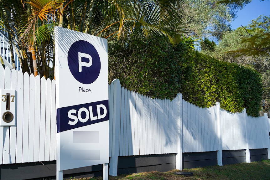 A 'sold' real estate sign sits next to a white fence outside a home