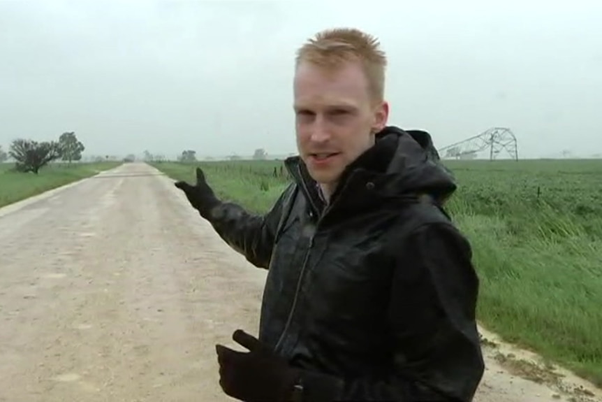 A reporter stands on a gravel road with a fallen transmission tower in the distance behind him.