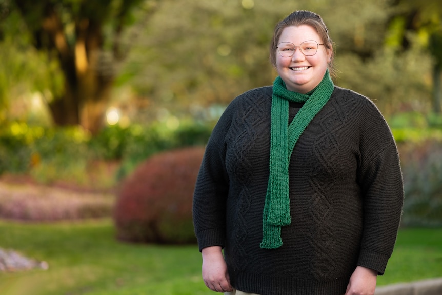 A woman in glasses with a green scarf stands in a park.