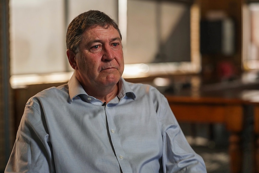 Man in blue button up shirt sits in front of camera. His eyes look teary