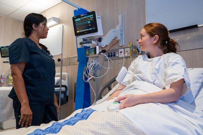 A patient resting in a hospital bed, a health worker is standing next to her and looking at a health monitoring screen nearby.