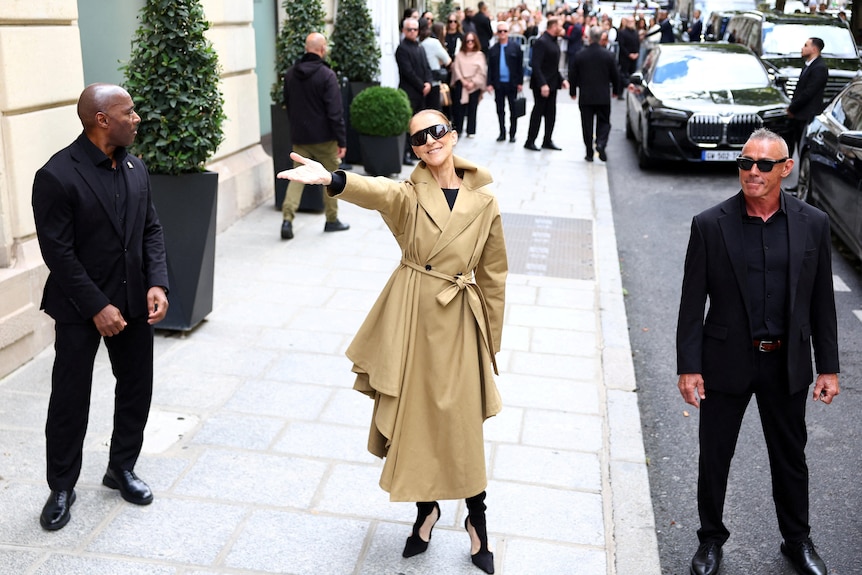 Celine Dion greets fans outside her hotel in Paris. She wears a trench coat and glasses