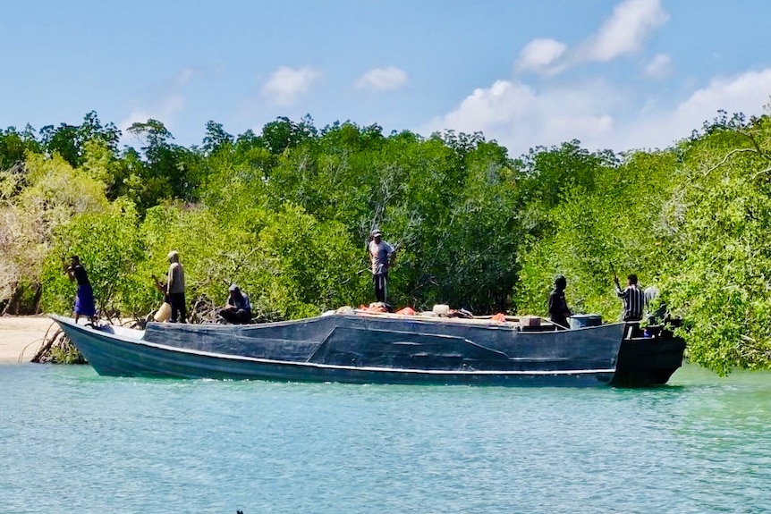 A boat in shallow water in front of green vegetation with a handful of men onboard
