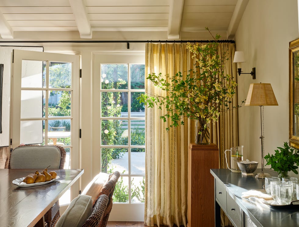 Bright interior with open French doors, a dining table, potted plants, and warm light filtering through curtains.