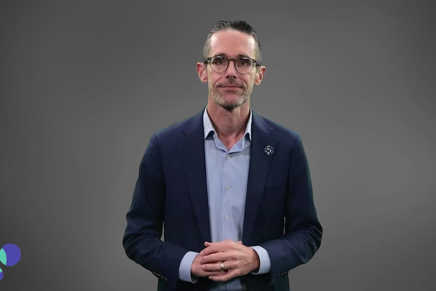 A man in a suit jacket and glasses with hands folded looking into camera on grey background.