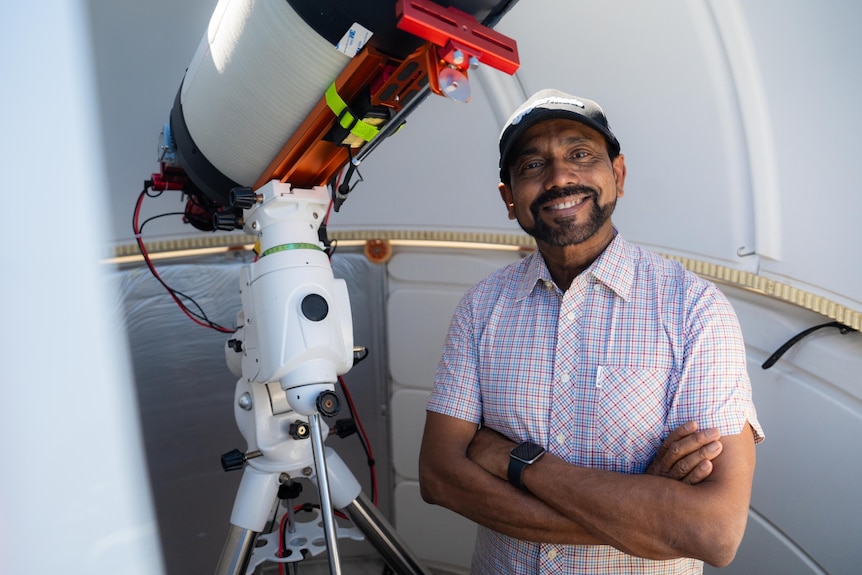 A man wearing a cap smiling and standing next to a telescope.
