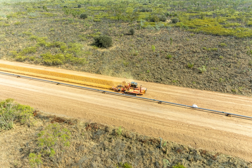 Aerial shot of yellow grader machine moving across dirt surface, with gray pipeline visible from left to right