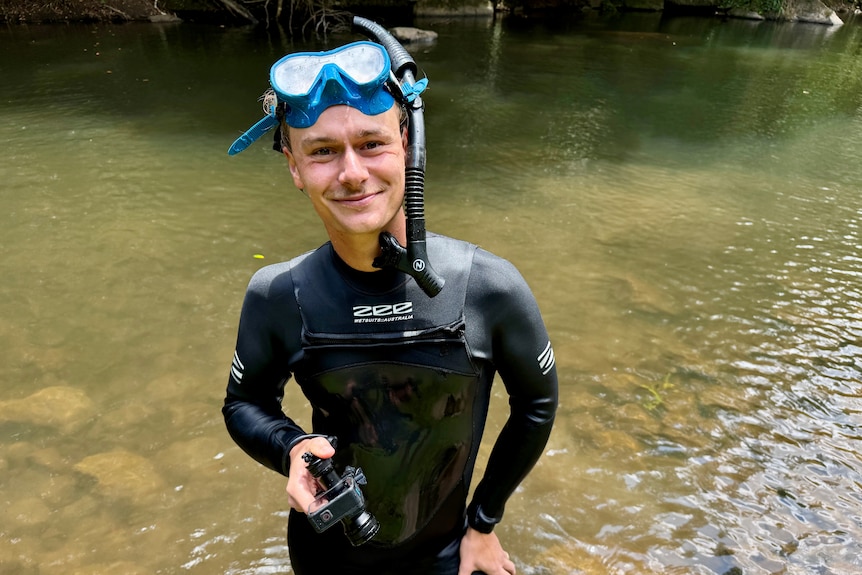 A man in a wetsuit with a Go-Pro and scuba mask smiles at the camera from a creek.
