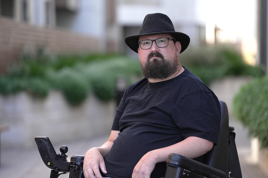 A white adult man with glasses and a black brimmed hat sitting in a wheelchair