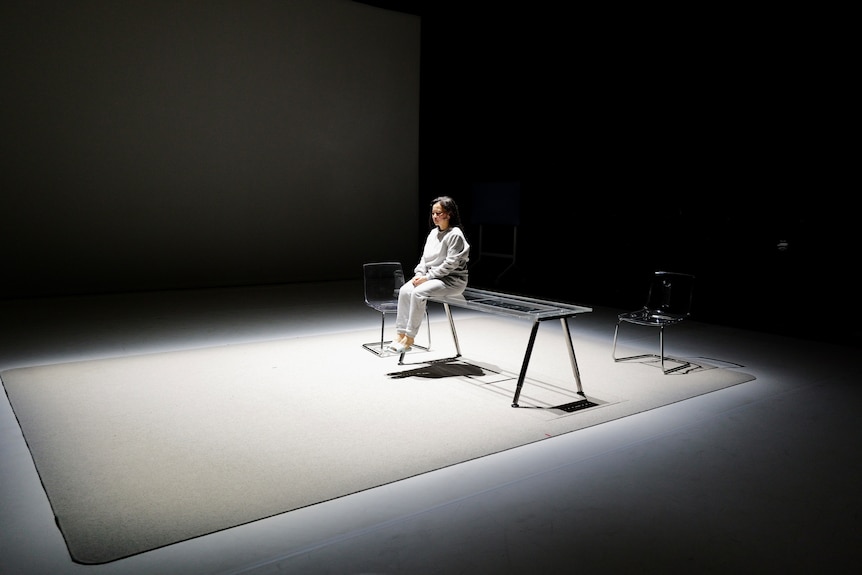 Cheng Lei acts on stage in her play. She sits alone on a desk in an empty space. 