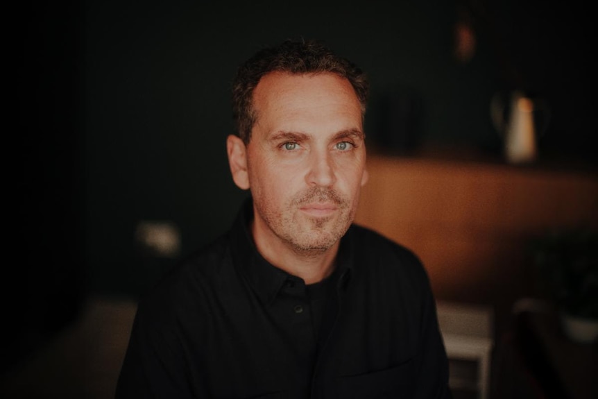 Dark-haired man in his late '40s with grey facial hair poses indoors in a black collared shirt