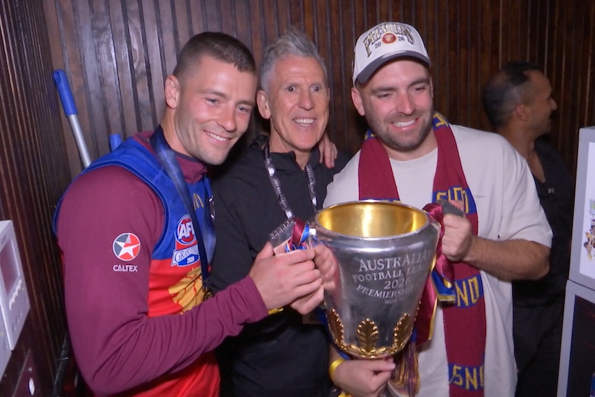 Three men, some dressed in Brisbane Lions gear, stand arm in arm holding a big silver cup.