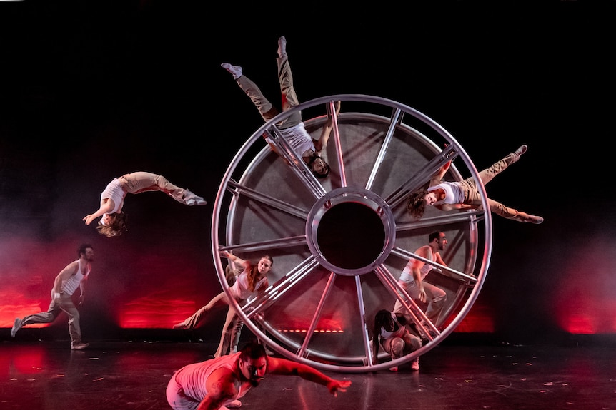 Acrobats performing with a giant wheel onstage in front of black background.