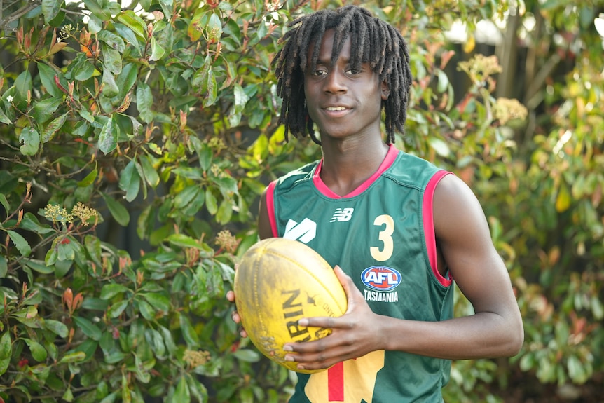 A young man wearing a green guernsey holding a yellow football and smiling.