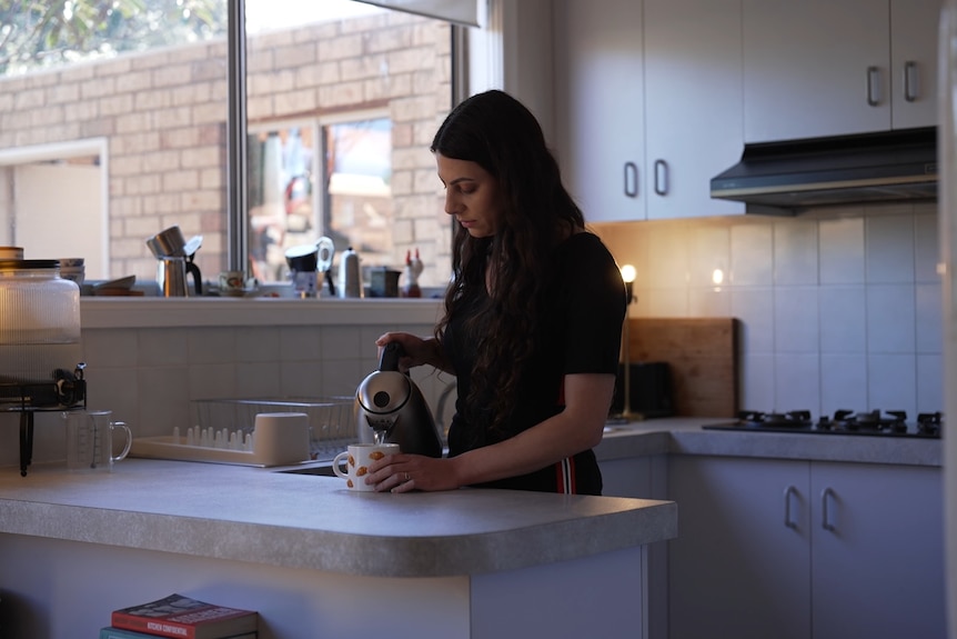 A dark-haired woman fills a mug ffrom a kettle while standing in a kitchen.